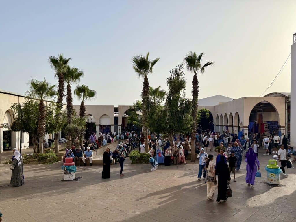 Vista general del Souk El Had de Agadir al atardecer, con palmeras y decenas de personas caminando y descansando en bancos, reflejando la diversidad social y estilística de Marruecos.
