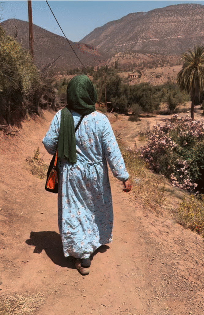 Mujer con pañuelo verde y caftán azul estampado de algodón caminando por un sendero rural cerca de Amzmiz, Marruecos