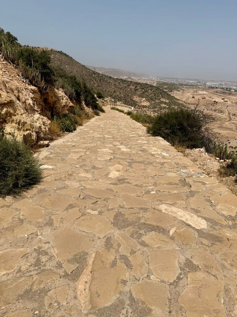 Camino empedrado de bajada desde la colina de Agadir Oufella entre vegetación y vistas al paisaje