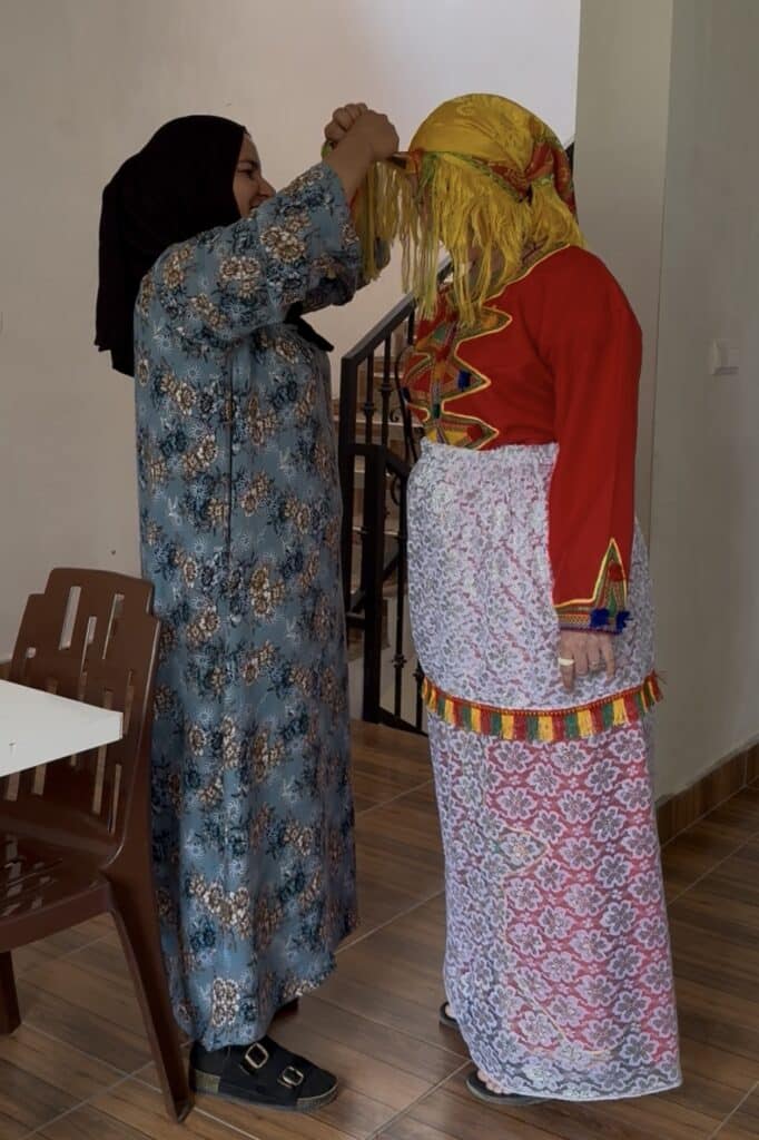 Mujer ayudando a colocar el tocado a otra vestida con traje tradicional amazigh rojo, encaje blanco y adornos de colores