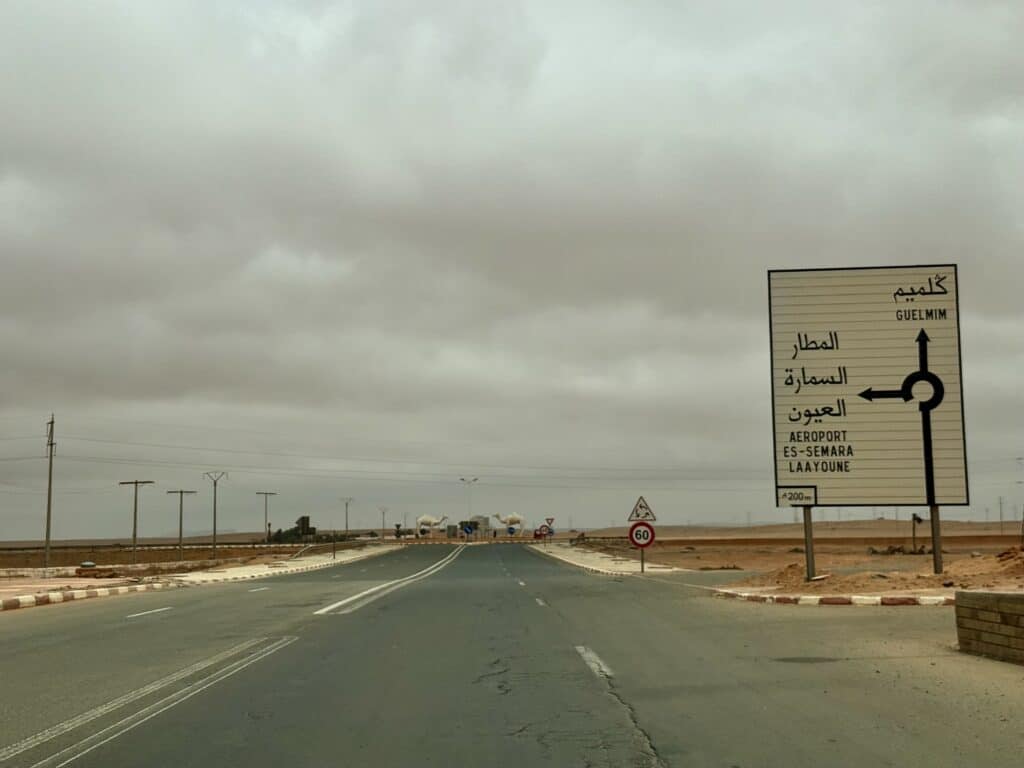 Cruce de carreteras en el sur de Marruecos con señalización hacia El Aaiún, Es-Semara y el aeropuerto, bajo un cielo nublado y tonos desérticos