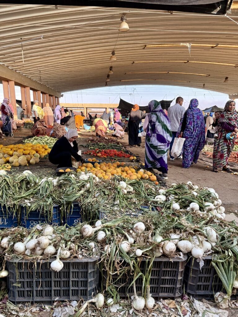 Mujeres con melhfa en tonos morados, verdes y azules caminan y venden productos en el mercado cubierto de Guelmim