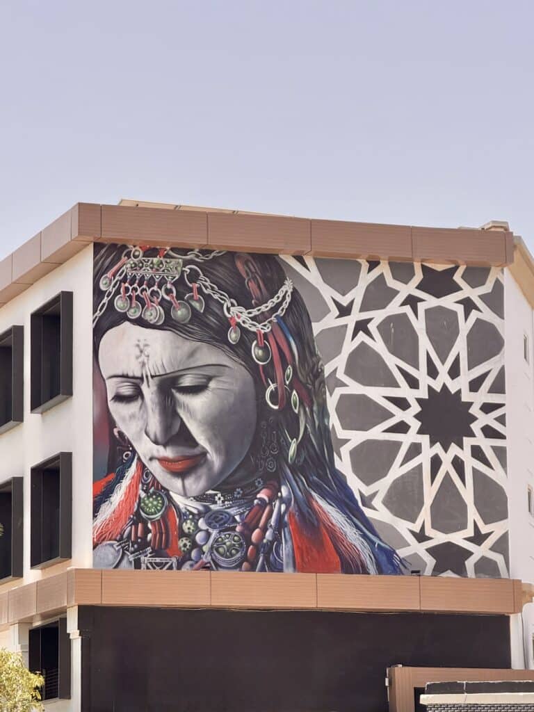 Mural urbano en la fachada de un edificio moderno que retrata a una mujer amazigh con tatuajes faciales tradicionales y joyería simbólica, destacando la fuerza de la identidad femenina y cultural