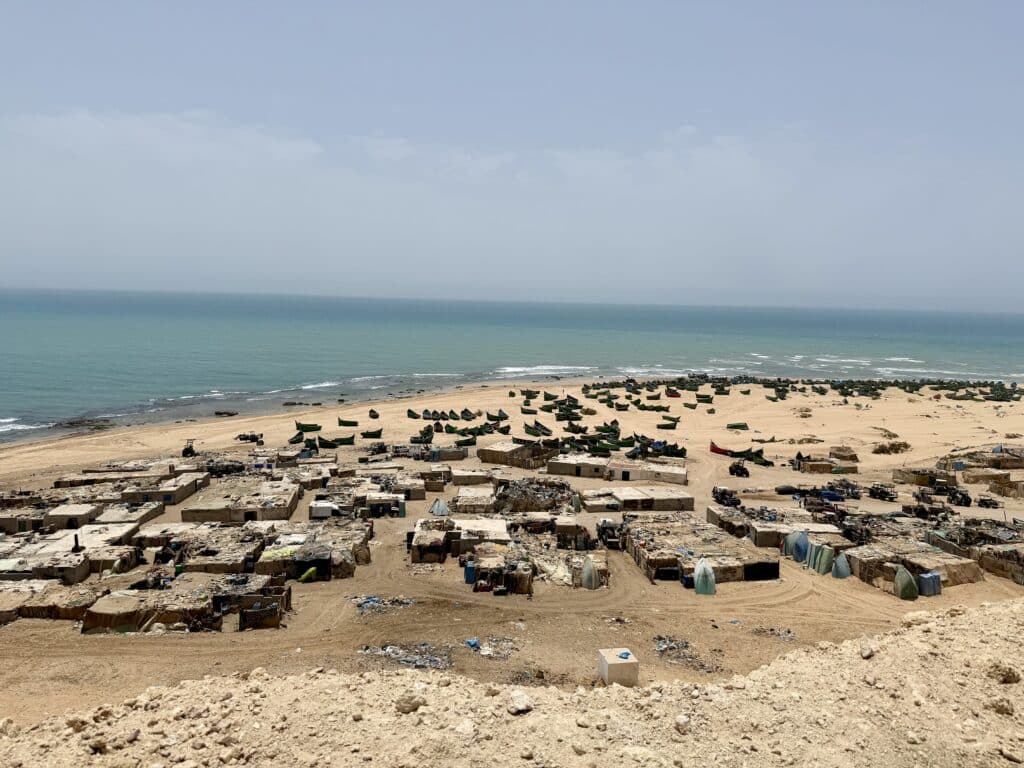 Vista panorámica de un asentamiento pesquero precario con barcas verdes sobre la arena y casas construidas con materiales reutilizados, en la costa del Sáhara Occidental