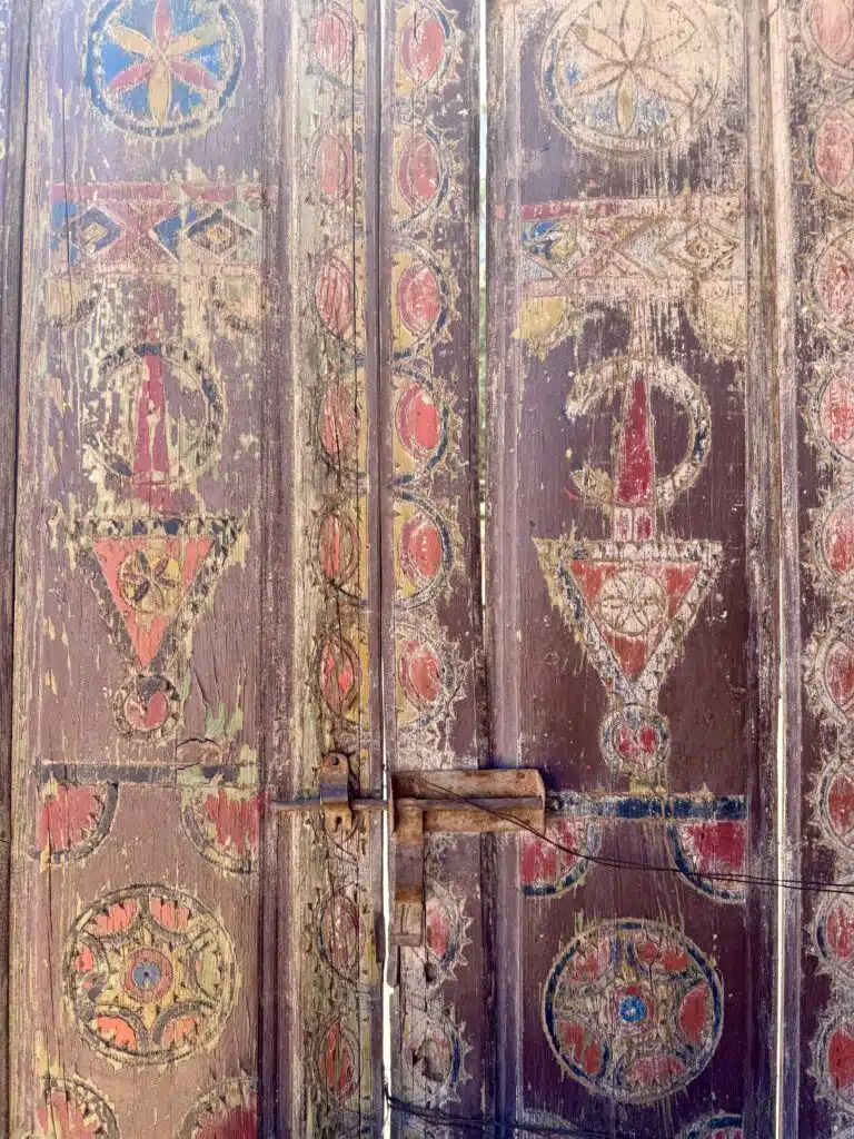 Puerta antigua de madera pintada con motivos amazigh en tonos rojos y amarillos, visiblemente desgastada por el tiempo.