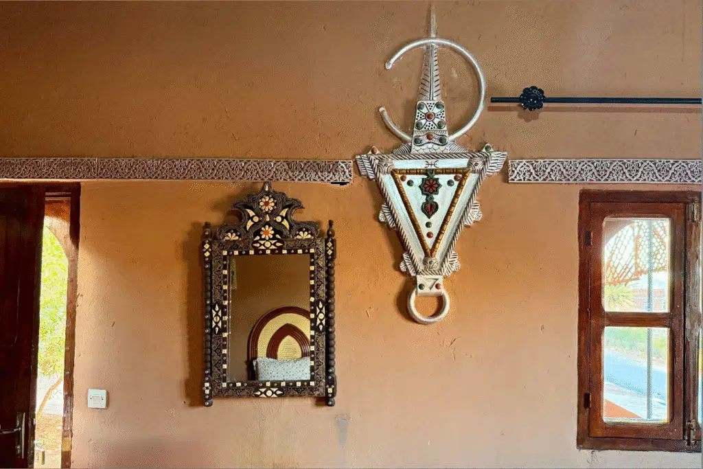 Interior de una casa tradicional marroquí con un espejo decorado y un gran colgante amazigh en la pared, junto a una ventana de madera.