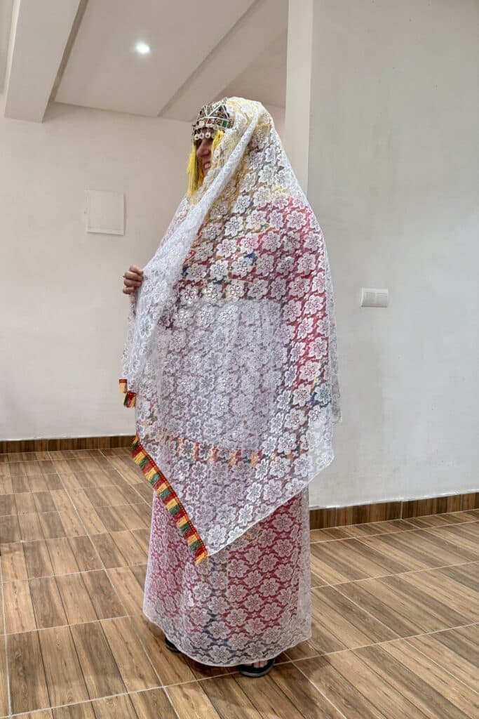 Mujer con vestido tradicional amazigh cubierto por un manto de encaje blanco con bordes de colores y joyería tradicional con monedas
