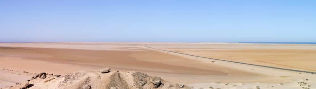 Vista panorámica desde una colina sobre el paisaje desértico y la carretera que bordea el Atlántico en el Sáhara Occidental, con tonos ocres y azul intenso del mar